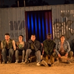 Seven actors sit in a row in front of an industrial background.