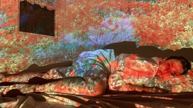 Young woman lays head on pillow with neutral expression. An image of a red flowers and other plants are projected over the scene.